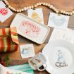 Christmas-themed decor on a wooden table, including a “Reindeer Feed” tray, snowman plate, bird mug, cards with festive designs, a wrapped gift, pine with berries, and a white bead garland.