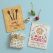 A wooden cutting board with baking utensils, a Milk & Cookie Co. holiday sign, small Christmas ornaments, and a wrapped gift are arranged on a light blue background.