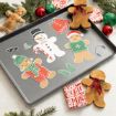 A baking tray with paper cutouts of decorated gingerbread people and winter accessories; real gingerbread cookies, presents, ornaments, and pine branches surround the tray on a marble surface.