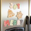 Colorful, festive magnets spelling “JOY” and “MERRY,” with a decorative bell and snowflake, are displayed on a stainless steel refrigerator door.