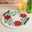 A round white tray with red poinsettias and green leaves, black handles, sits on a countertop near two lit candles and a small, decorated Christmas tree.
