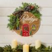 A festive wreath with greenery, berries, and a plaid bow hangs on a white wall. It reads “Home for the Holidays” and features a red door design. Three lit candles and pine branches are displayed below it.