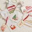 Three festive drawstring gift bags with Christmas designs and coordinating tags featuring Santa, a reindeer, and penguin, all set on a light, textured surface. Red and gold striped paper tags are also included in the arrangement.