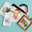 Christmas-themed items on a blue background: a towel with a reindeer, a towel with a snowman, a tote bag with a cow in a Santa hat, a gold-framed Santa picture, and pine branches with berries.