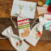 Three festive fabric gift bags with Christmas designs on a wooden table, surrounded by wrapping paper, tags, a wax seal, and a card with holiday ornaments.