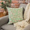 A holiday-themed pillow with a green and red holly pattern rests on a gray couch next to a cream knit blanket. A small decorated Christmas tree is in the background next to a brick wall.