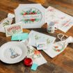 Assorted Christmas-themed items on a wooden floor, including a plate with a snowman, a mug, coasters, prints, two drawstring bags, a red candle, and “Reindeer Feed” cards, all with festive designs and illustrations.