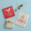 Flat lay of two festive signs, three Christmas ornaments, and a small wrapped gift with a gold ribbon, all arranged on a light blue background.
