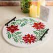 A round tray with painted red poinsettias and green leaves, black handles, sits on a countertop near two lit candles, a small Christmas tree, and a greeting card.