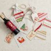 A bottle of wine with festive holiday gift bags, tags featuring Santa and a reindeer, and patterned note cards arranged on a light surface.