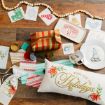 Holiday-themed items on wood floor: greeting cards, red-green wrapped gift, festive pillow, mug, plate, tray, ribbon, string of wooden beads, candle, pouch, and gift tags with holiday prints.