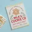 A sign with a gold snowflake reads, “Original North Pole Milk & Cookie Co. Baking Spirits Bright. Gingerbread, Cakes, Cookies.” A fabric with script text is partially visible on the left.