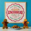 A sign reads “Old Fashioned Gingerbread Cookie Co. Baked Fresh Daily.” Two gingerbread figures with bows stand beside a plate of cookies, all set against a blue wall.
