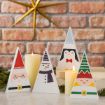 Four triangular wooden figures painted as Santa, a snowman, a penguin, and an elf are arranged with two lit candles in front of a brick wall and a gold star decoration.