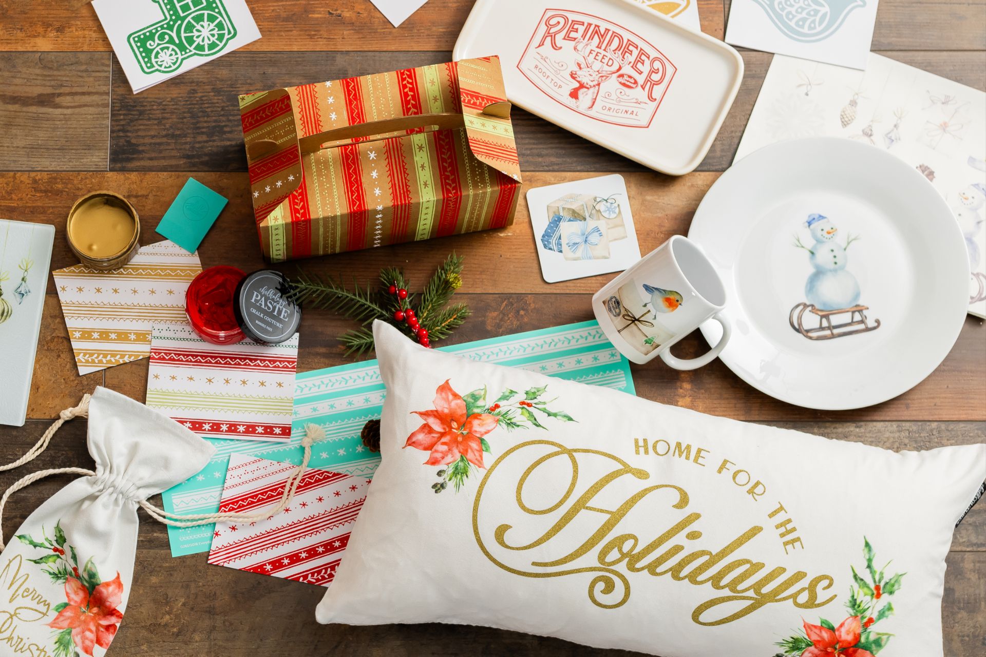 Assorted holiday-themed items on a wooden table, including a pillow with “Home for the Holidays,” a snowman plate, a reindeer tray, a wrapped gift, festive cards, a mug, ribbon, a small gift bag, and a candle.