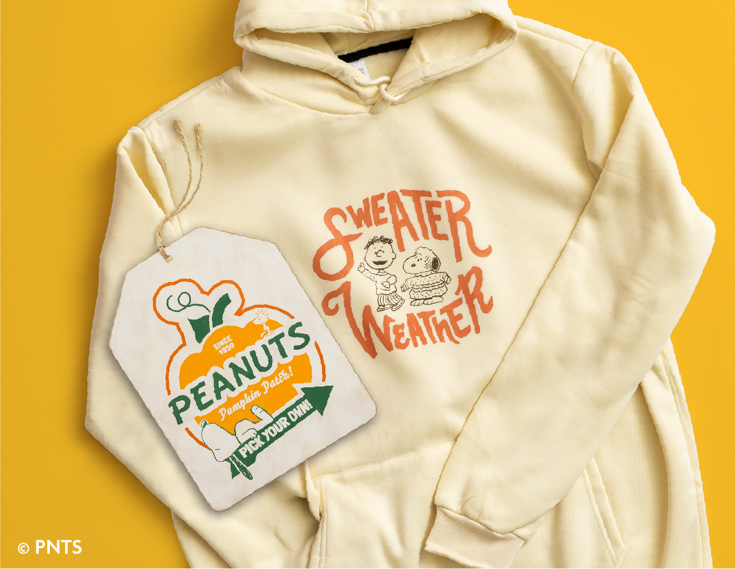 A cream hoodie with a “Sweater Weather” graphic featuring Peanuts characters. Next to it is a pumpkin-shaped tag reading “Peanuts Pumpkin Patch, Pick Your Own” on a yellow background.