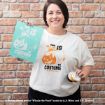 Smiling woman in white shirt with an orange Pooh bear “This is my costume” design, holding matching art print and a small round item, stands against a brick wall.