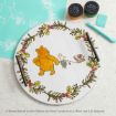 A round wooden tray with handles features Winnie the Pooh and Piglet pulling a small cart, surrounded by a painted wreath of leaves and red berries. Craft supplies and a stencil are visible nearby.