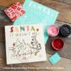 A wooden block with “Santa, It Wasn’t Me!” and a sketch of Winnie the Pooh and Tigger, surrounded by red and pink craft supplies, wrapping paper, and a blue card.
