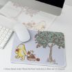 A mouse pad with Winnie the Pooh and Piglet collecting apples from a tree, next to a white mouse and keyboard on a desk.