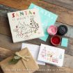 Flat lay of Christmas cards, an envelope, a brown gift, red and pink ink, and a box with Winnie the Pooh art on a wooden table. Text includes “SANTA, IT WASN’T ME!” and “MERRY EVERYTHING.”.