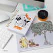 A desk setup with a Disney Winnie the Pooh notebook, a mouse pad showing Pooh and Piglet by an apple tree, a keyboard, a mouse, a blue coaster, and a black and yellow jar.