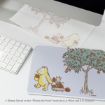 A mouse pad and desk mat featuring Winnie the Pooh and Piglet holding hands near an apple tree with apples scattered on the ground, placed beside a computer keyboard.