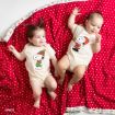 Two babies lie on a red polka dot blanket. Both wear cream-colored onesies with Snoopy holiday designs—one as an elf, the other as Santa. They appear happy and are looking in different directions.