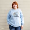 A smiling woman with red hair wears a light blue “Chasing Snowflakes” Snoopy sweatshirt and jeans, standing against a beige wooden wall with one hand in her pocket.