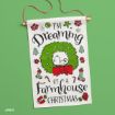 A festive wall hanging with a dog inside a green wreath reads: “I’m dreaming of a farmhouse Christmas.” Decorated with gifts, stockings, snowflakes, holly, and trees, on a green background.