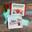 Home for the holidays sign with Peanuts characters, paint jars, a blue print, and framed A Charlie Brown Christmas artwork on a wooden surface.