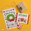 A farmhouse Christmas wall hanging, two wrapped gifts, and a holiday card with Peanuts characters sit on a yellow background. The card reads “Home for the Holidays!” and the hanging says “I’m Dreaming of a Farmhouse Christmas.”.