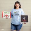 A smiling woman in a blue “Chasing Snowflakes” sweatshirt holds a “Home for the Holidays!” sign and a “Reindeer Crossing” framed decoration against a light wood wall.
