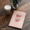 A spiral notebook with a coffee cup illustration, a pencil, and a takeaway coffee cup sit on a wooden table next to a vase of purple flowers.