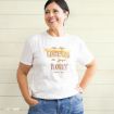Smiling woman in a white t-shirt with the text “I’m the Lorelai to your Rory” stands with one hand on her hip against a light wooden background.