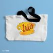 A white tote bag with black straps features an orange coffee cup and the word “Luke’s” in bold letters on the front, photographed on a light blue background.