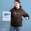 A smiling woman in a brown hoodie that says “Sip coffee, read books, Gilmore Girls” holds a Chilton Preparatory School sign against a blue wall, pointing at her sweatshirt.