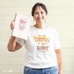 Smiling woman holding a pink notebook with a coffee cup illustration, wearing a white shirt that says “I’m the Lorelai to your Rory.”.