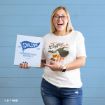A smiling person in glasses and jeans holds a Chilton Preparatory School folder and points to their shirt, which says “I need coffee in an IV,” with coffee graphics, standing against a blue wooden wall.