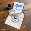 A tote bag with “Life’s Short, Talk Fast Gilmore girls” printed on it sits on a wooden floor next to a coffee cup and a Chilton preparatory school folder with flowers tucked inside.