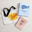 A white tote bag with “Luke’s” in yellow and orange, a blue “Chilton” folder, and a pink spiral notebook with a coffee cup illustration, all arranged on a white surface.