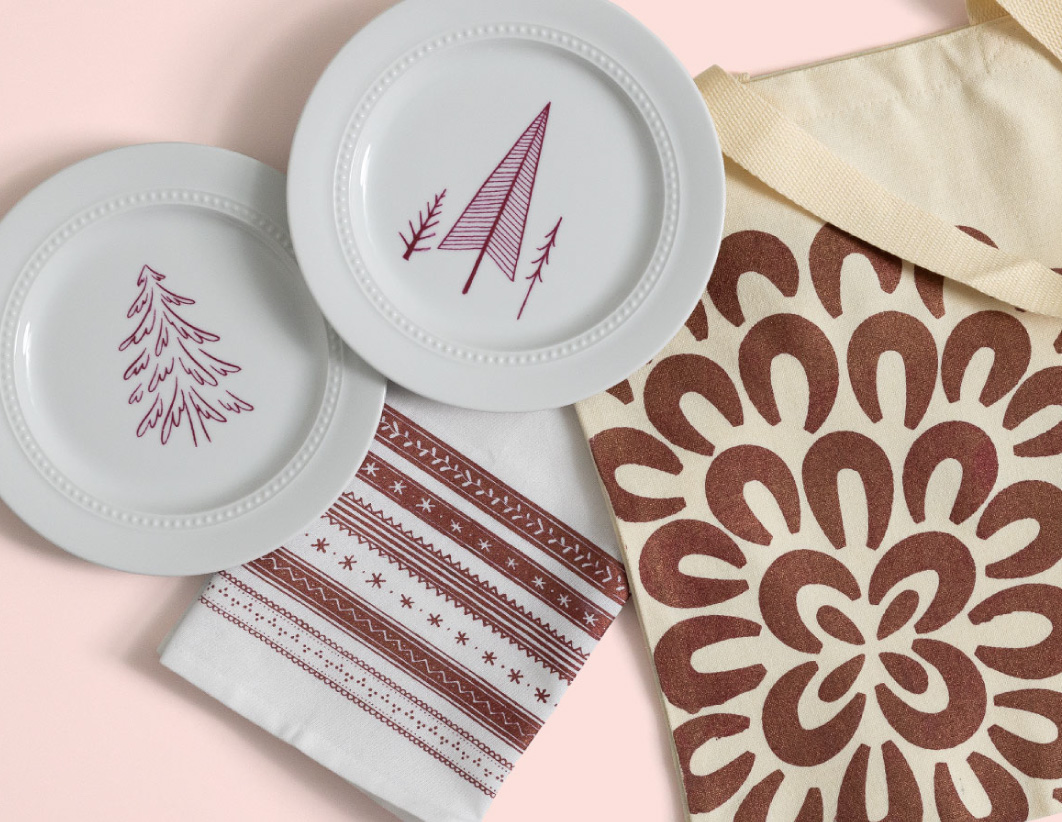 Two white plates with red tree designs, a matching napkin with red patterns, and a cream tote bag with a bold brown floral print are arranged on a pale pink background.