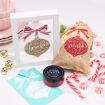 Holiday-themed items, including a framed ornament print, a decorated gift bag, candy canes, mint candies, a chalk paste jar, a stencil, and stacked pink dishes, are displayed on a white surface.