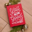 A red sign with white festive lettering reads “Y’all be good now” beside gold jingle bells and evergreen sprigs, on a wooden surface.