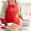 A person in a red “Mrs. Claus’ Christmas Kitchen” apron holds a tray of cookies and a recipe card in a bright kitchen.