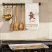Wooden spoons and measuring cups hang above a stove. A towel with a broken gingerbread man and “Oh Snap!” text is draped on the rack. A baking tray with cookies sits on the stove below.
