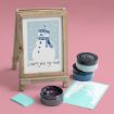 A framed snowman illustration with the text “I CAN’T FEEL MY TOES!” sits on a small easel. Nearby are jars of chalk paste, a mint green tool, and a matching snowman card, all on a pink surface.