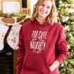 Smiling woman in a red hoodie that says “Too cute for the naughty list” stands in front of a decorated Christmas tree and stockings hanging on a fireplace mantel.