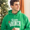 A smiling young man in a green “Team Grinch” hoodie holds a mug while standing near a decorated Christmas tree.