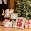 Christmas gifts, framed Santa art, and decorations sit by a lit fireplace and a decorated Christmas tree with gold and red ornaments. Cozy blankets and holiday pillows are on a nearby chair.